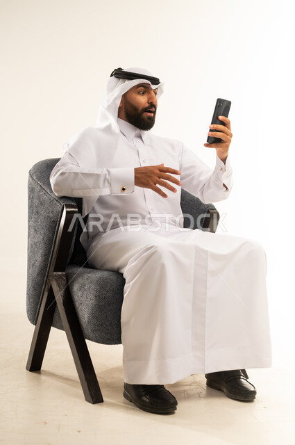 ايماءات الصدمة والذهول، دمج التكنولوجيا والاجهزة الحديثة في الحياة اليومية، تصفح مواقع التواصل الاجتماعي عبر الانترنت، بورتريه لرجل عربي خليجي قطري مبتسم يرتدي الثوب والغترة يجلس على كرسي مريح يقوم باستخدام الهاتف المحمول بتعابير الاعجاب والدهشة من شيء ما، خلفية بيضاء
