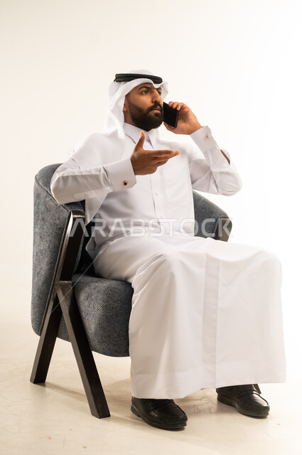 Benefiting from mobile services and applications, integrating technology and modern devices into daily life, portrait of a Qatari Gulf Arab man wearing traditional thobe and ghutra sitting on a comfortable chair making a phone call using a mobile phone, communicating with family and friends, chatting, conversations and getting to know each other, white background