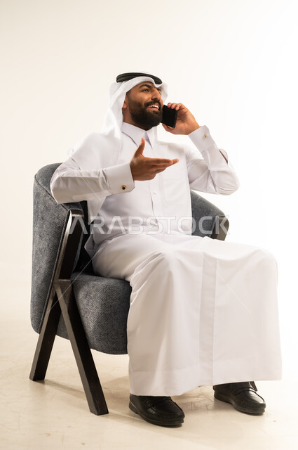 Benefiting from mobile services and applications, integrating technology and modern devices into daily life, portrait of a Qatari Gulf Arab man wearing traditional thobe and ghutra sitting on a comfortable chair making a phone call using a mobile phone, communicating with family and friends, chatting, conversations and getting to know each other, white background