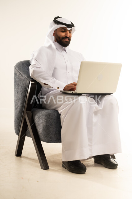 مفهوم التوظيف أونلاين، تصفح مواقع التواصل الاجتماعي باستخدام الحاسوب المحمول، العمل عن بعد عبر الانترنت، دمج التقنيات الحديثة والتكنولوجيا في الحباة العملية, بورتريه لرجل عربي خليجي قطري يرتدي الثوب التقليدي والغترة يجلس على كرسي مريح يقوم بالنقر على لوحة مفاتيح اللابتوب، خلفية بيضاء
