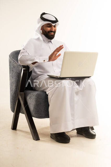 ايماءات الترحيب والتحية والسلام، دمج التقنيات الحديثة والتكنولوجيا في الحياة اليومية، التواصل مع الاهل والاصدقاء عبر الانترنت باستخدام الحاسوب المحمول, بورتريه لرجل عربي خليجي قطري يرتدي الثوب التقليدي والغترة يجلس على كرسي مريح يقوم بإجراء مكالمة فيديو عبر جهاز اللابتوب، خلفية بيضاء