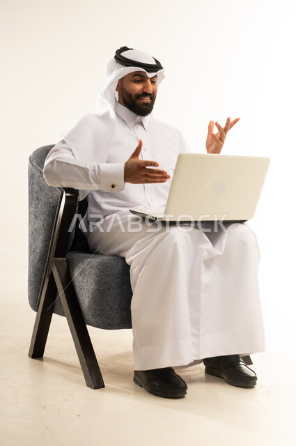 مفهوم التوظيف أونلاين، دمج التقنيات الحديثة والتكنولوجيا في الحياة العملية، العمل عن بعد عبر الانترنت باستخدام الحاسوب المحمول, بورتريه لرجل عربي خليجي قطري يرتدي الثوب التقليدي والغترة يجلس على كرسي مريح يقوم بإجراء مقابلة عمل عبر جهاز اللابتوب، خلفية بيضاء