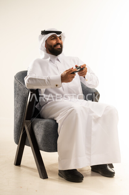 قضاء اوقات ممتعة بألعاب الفيديو الترفيهية في عطلة نهاية الاسبوع، بورتريه لرجل عربي خليجي قطري يرتدي الثوب والغترة يجلس على كرسي مريح يمسك بيده عصا التحكم بتعابير الاندماج والسرور، اللعب والاستمتاع بأوقات الفراغ، دمج التقنية والتكنولوجيا في الحياة اليومية، خلفية بيضاء