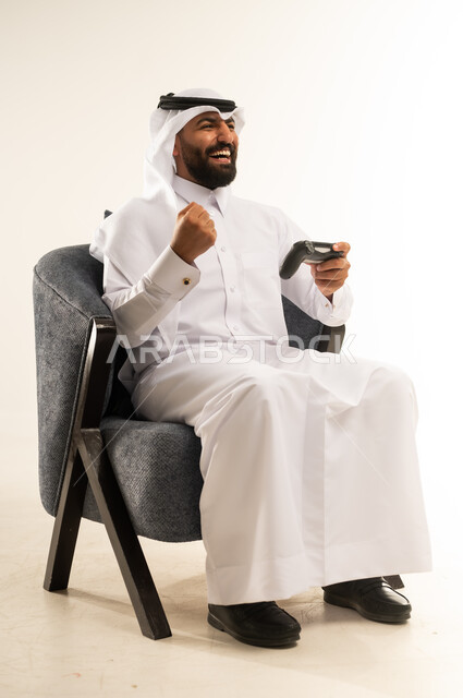 مفهوم الانجاز وتحقيق الاهداف، الاندماج والتفاعل في ألعاب الفيديو الترفيهية، بورتريه لرجل عربي خليجي قطري مبتسم يرتدي الثوب والغترة يجلس على كرسي مريح يمسك بيده عصا التحكم يقوم برفع قبضة يده بتعابير الفوز والنجاح، دمج التقنية والتكنولوجيا في الحياة اليومية، خلفية بيضاء