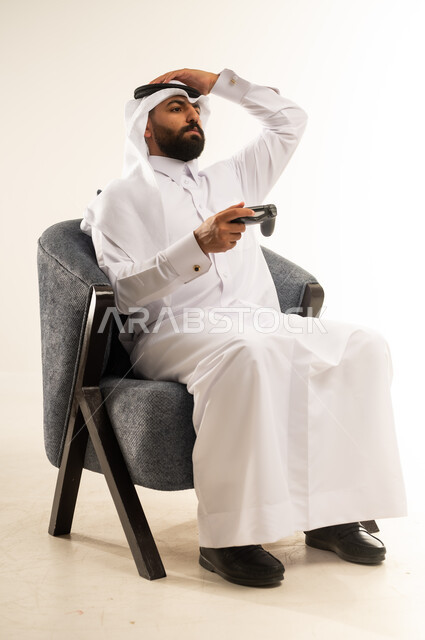 الاندماج والتفاعل في ألعاب الفيديو الترفيهية، بورتريه لرجل عربي خليجي قطري مبتسم يرتدي الثوب والغترة يجلس على كرسي مريح يمسك عصا التحكم يقوم بوضع يده عبى رأسه بتعابير الخسارة والهزيمة والانزعاج ، دمج التقنية والتكنولوجيا في الحياة اليومية، خلفية بيضاء