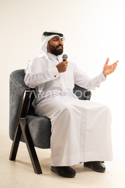 ايماءات الاستمتاع والتفاعل والاندماج مع الانغام الموسيقية ، تنمية المواهب الصوتية والغنائية، بورتريه لرجل عربي خليجي قطري مبتسم يرتدي الثوب التقليدي والغترة يجلس على كرسي مريح يقوم بتسجيل مقطع بصوتي باستخدام مكبر الصوت ، خلفية بيضاء