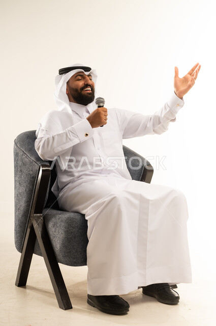 ايماءات الاستمتاع والتفاعل والاندماج مع الانغام الموسيقية ، تنمية المواهب الصوتية والغنائية، بورتريه لرجل عربي خليجي قطري مبتسم يرتدي الثوب التقليدي والغترة يجلس على كرسي مريح يقوم بتسجيل مقطع بصوتي باستخدام مكبر الصوت ، خلفية بيضاء