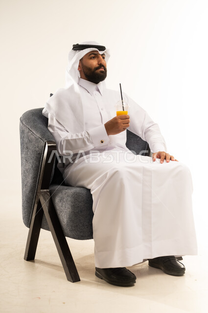 الحصول على كأس من العصير البارد والمنعش، الجلوس ومحاولة الراحة والاستمتاع بأوقات الفراغ، بورتريه  لشاب عربي خليجي قطري يرتدي الثوب التقليدي والغترة يجلس على كرسي مريح يمسك بيده كوب من المشروب الصيفي المفضل، خلفية بيضاء