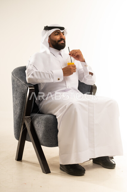 الحصول على كأس من العصير البارد والمنعش، الجلوس ومحاولة الراحة والاستمتاع بأوقات الفراغ، بورتريه  لشاب عربي خليجي قطري يرتدي الثوب التقليدي والغترة يجلس على كرسي مريح يمسك بيده كوب من المشروب الصيفي المفضل، خلفية بيضاء