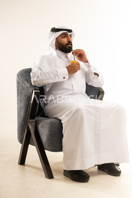 الحصول على كأس من العصير البارد والمنعش، الجلوس ومحاولة الراحة والاستمتاع بأوقات الفراغ، بورتريه  لشاب عربي خليجي قطري يرتدي الثوب التقليدي والغترة يجلس على كرسي مريح يمسك بيده كوب من المشروب الصيفي المفضل، خلفية بيضاء