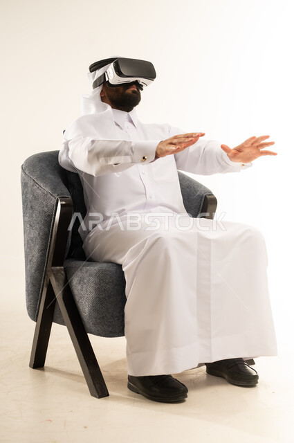 الاندماج والتفاعل ومحاكاة العالم الخيالي، بورتريه لرجل عربي خليجي قطري يرتدي الثوب التقليدي والغترة ونظارات الواقع الافتراضي يجلس على كرسي مريح يقوم بمد يديه الى الامام بتعابير محاولة لمس شيء ما، دمج التسلية مع التكنولوجيا والتقنيات الحديثة، خلفية بيضاء