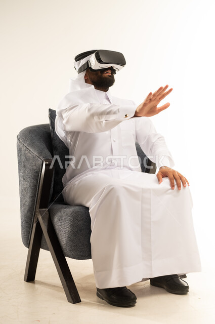 المتعة والتسلية  بالمشاهدة عبر نظارة الواقع الافتراضي, بورتريه لرجل عربي خليجي قطري يرتدي الثوب التقليدي والغترة يجلس على اريكة مريحة يضع نظارات ثلاثية الأبعاد يقوم برفع وفتح كف يده للاعلى بتعابير الاندماج والتوقف عن فعل امر ما, خلفية بيضاء