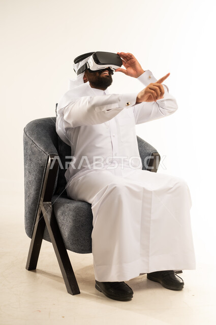 محاولة النقر على شيء ما, المتعة والتسلية  بالمشاهدة عبر نظارة الواقع الافتراضي, بورتريه لرجل عربي خليجي قطري يرتدي الثوب التقليدي والغترة يجلس على اريكة مريحة يضع نظارات ثلاثية الأبعاد يقوم برفع اصبع السبابة للاعلى والاشارة الى مكان ما بتعابير الاندماج ومحاكاة العالم الخيالي, خلفية بيضاء