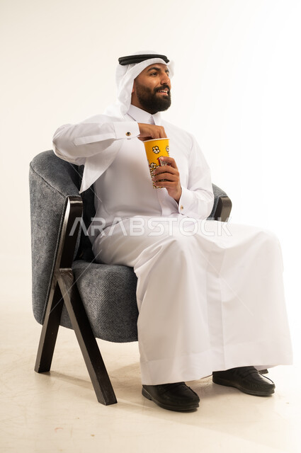 الجلوس ومشاهدة الأفلام في أوقات الفراغ، إيماءات الاندماج والتركيز مع الفيلم، الاستمتاع بتناول الفشار، بورتريه لشاب عربي خليجي قطري يرتدي الثوب التقليدي والغترة يجلس على كرسي مريح ويمسك بيده علبة من البوب كورن اللذيذ، خلفية بيضاء