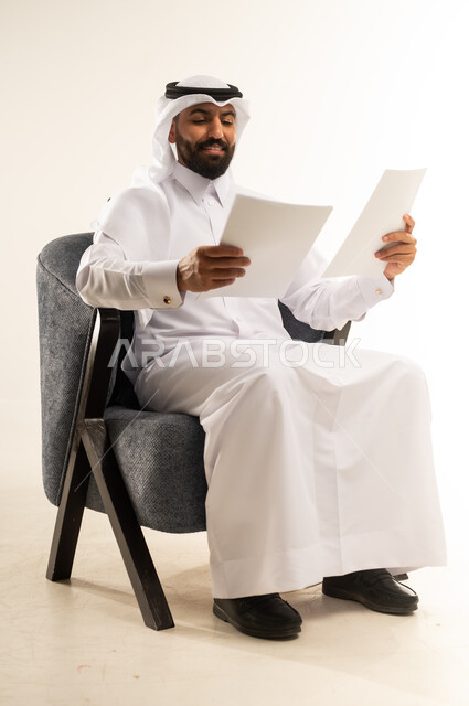 Following up on plans and projects, reading notes and information, portrait of a Qatari Gulf Arab man wearing traditional thobe and ghutra sitting on a comfortable chair reading and examining papers with expressions of concentration and contemplation, concept of getting work done, white background
