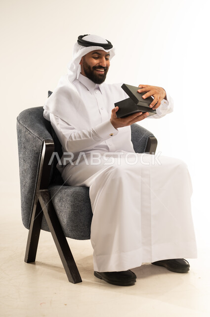 مفهوم المفاجأة والإهداء، إيماءات السعادة والدهشة بالحصول على هدية ثمينة، فن انتقاء الهدايا بعناية، بورتريه لشاب عربي خليجي قطري مبتسم  يرتدي الثوب التقليدي والغترة يجلس على كرسي ويقوم بفتح العلبة بتعابير الفرح والسرور، خلفية بيضاء