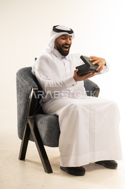 مفهوم المفاجأة والإهداء، إيماءات السعادة والدهشة بالحصول على هدية ثمينة، فن انتقاء الهدايا بعناية، بورتريه لشاب عربي خليجي قطري مبتسم  يرتدي الثوب التقليدي والغترة يجلس على كرسي ويقوم بفتح العلبة بتعابير الصدمة، خلفية بيضاء
