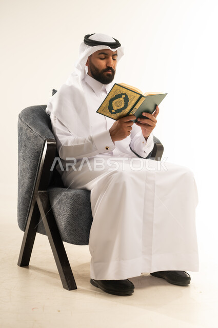 حفظ وتفسير كلام الله عز وجل، أداء العبادات والتقرب إلى الله تعالى، التعبد والتضرع بخشوع، بورتريه  لشاب عربي خليجي قطري يرتدي الثوب التقليدي والغترة يجلس على كرسي مريح يقوم بتلاوة وترتيل القرآن الكريم, خلفية بيضاء