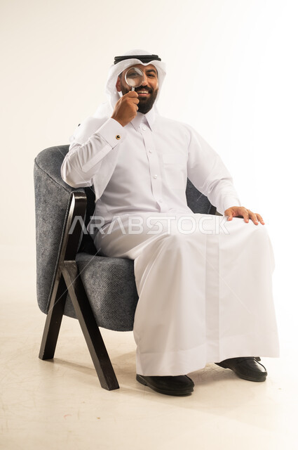 تكبير وتوضيح الرؤية ، عمليات البحث والتدقيق، مضاعفة الأحجام ومحاولة إيجاد الأشياء الصغيرة، بورتريه لشاب عربي خليجي قطري يرتدي الثوب التقليدي والغترة يجلس على كرسي يحاول اكتشاف شيء ما باستخدام العدسة المكبرة بإيماءات الاندماج والسرور، خلفية بيضاء