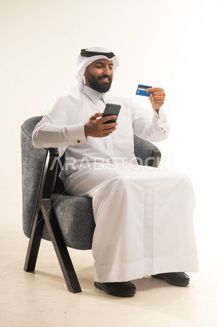 مفهوم التسوق أونلاين، عمليات التبضع والشراء عبر تطبيقات الانترنت، استخدام التقنيات الحديثة والتكنولوجيا, بورتريه لرجل عربي خليجي قطري يرتدي الثوب التقليدي والغترة يجلس على كرسي مريح يقوم بعملية الدفع الالكتروني بالاعتماد على البطاقة البنكية والجوال، خلفية بيضاء