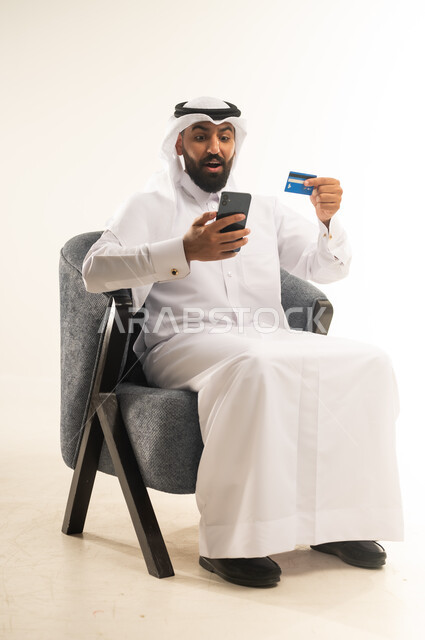 مفهوم التسوق أونلاين، عمليات التبضع والشراء عبر تطبيقات الانترنت، استخدام التقنيات الحديثة والتكنولوجيا, بورتريه لرجل عربي خليجي قطري يرتدي الثوب التقليدي والغترة يجلس على كرسي مريح يقوم بعملية الدفع الالكتروني بالاعتماد على البطاقة البنكية والجوال، خلفية بيضاء