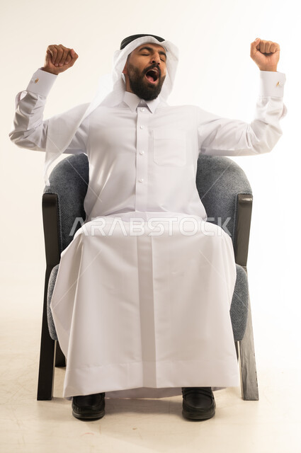 الشعور بالتعب والحاجة الى النوم والراحة, الإحساس بالكسل والخمول، بورتريه  لرجل عربي خليجي قطري يرتدي الثوب التقليدي والغترة يجلس على كرسي مريح يقوم بتمديد جسمه ورفع ذراعيه للاعلى بإيماءات النعاس والتثاوب والارهاق, خلفية بيضاء