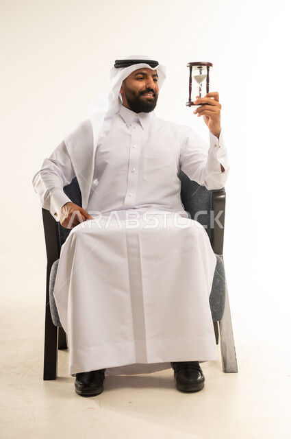 إدراك قيمة اللحظات الثمينة، مراقبة توقيت الرمل، مفهوم إدارة الأعمال وتنظيم الجدول الزمني, بورتريه لرجل عربي خليجي قطري مبتسم يرتدي الثوب التقليدي والغترة يمسك بيده الساعة الرملية الزجاجية (البنكام) بإيماءات الفرح والسرور، خلفية بيضاء