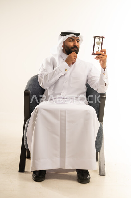 مراقبة توقيت الرمل، مفهوم إدارة الأعمال وتنظيم الجدول الزمني, إدراك قيمة اللحظات الثمينة, بورتريه لرجل عربي خليجي قطري يرتدي الثوب التقليدي والغترة يمسك بيده الساعة الرملية الزجاجية (البنكام) ويضع يده خلف ذقنه بإيماءات التفكير والتمعن، خلفية بيضاء