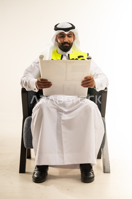Concept of engineering and architectural construction, engineering sector work, portrait of a Qatari Gulf Arab man wearing a protective vest and a ghutra sitting on a comfortable chair examining and reviewing construction plans with expressions of focus and contemplation, project management and follow-up, white background