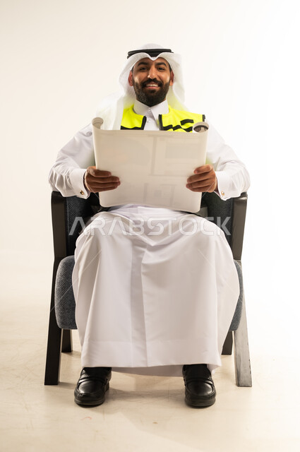 العمل في القطاع الهندسي، بورتريه لرجل عربي خليجي قطري يرتدي الثوب مع سترة الحماية والغترة يجلس على كرسي مريح يمسك مخططات الإعمار بيديه، النظر الى الكاميرا بتعابير الفخر والسرور، إدارة ومتابعة المشروع, مفهوم الهندسة والانشاءات المعمارية, خلفية بيضاء