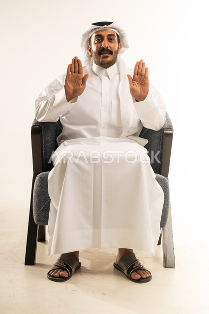 مفهوم الاناقة والاهتمام بالمظهر الخارجي، عرض ظاهر الكف والنظر الى الكاميرا بتعابير الفرح والسرور، بورتريه لرجل عربي خليجي قطري مبتسم يرتدي الثوب والغترة يجلس على كرسي مريح يرفع كفيه مفتوحة للاعلى، خلفية بيضاء