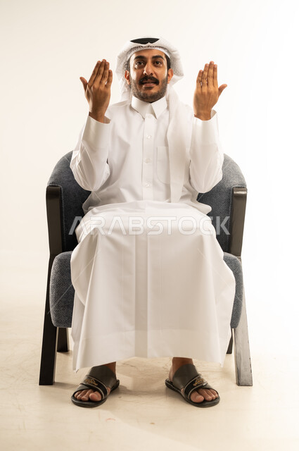 عرض باطن الكف والنظر الى الكاميرا بتعابير الفرح والسرور، بورتريه لرجل عربي خليجي قطري مبتسم يرتدي الثوب والغترة يجلس على كرسي مريح يرفع كفيه مفتوحة للاعلى، خلفية بيضاء