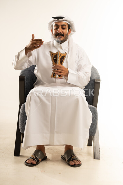 Following authentic Arab customs and traditions, incense and the use of perfumes and fragrances, incense burning on holidays and happy occasions, side portrait of a Qatari Gulf Arab man wearing traditional thobe and ghutra sitting on a chair and holding an incense burner in his hand, white background