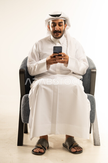 Communicating with people via mobile phone, chatting, conversations and getting to know each other, portrait of a Qatari Gulf Arab man wearing traditional thobe and ghutra sitting on a comfortable sofa using a mobile phone, browsing the internet and online shopping, using modern technologies and devices, white background