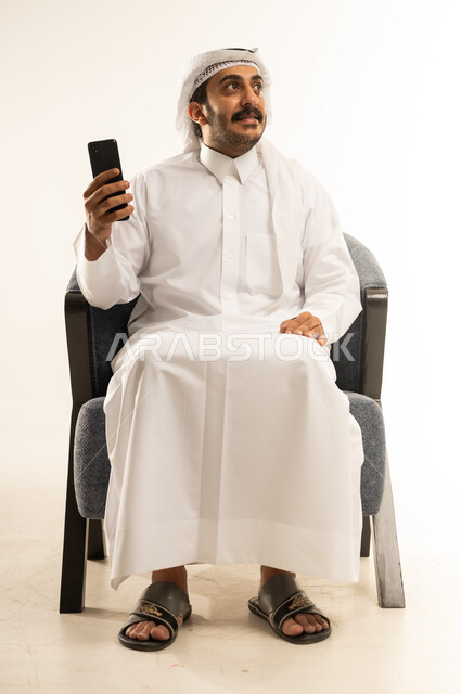 دردشة ومحادثات وتعارف، بورتريه لرجل عربي خليجي قطري يرتدي الثوب التقليدي والغترة يجلس على كنبة مريحة يستعمل الجوال، تصفح الانترنت والتسوق الالكتروني, استخدام التقنيات الحديثة، التواصل مع الافراد عبر الهاتف المحمول، النظر الى شيء ما بايماءات السرور، خلفية بيضاء