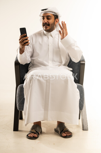ايماءات الترحيب والتحية والسلام، دمج التقنيات الحديثة والتكنولوجيا في الحياة اليومية، التواصل مع الاهل والاصدقاء عبر الانترنت , بورتريه لرجل عربي خليجي قطري يرتدي الثوب التقليدي والغترة يجلس على كرسي مريح يقوم بإجراء مكالمة فيديو عبر جهاز اللابتوب، خلفية بيضاء