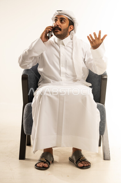 Expressions remembering something, portrait of a smiling Qatari Gulf Arab man wearing traditional thobe and ghutra sitting on a comfortable chair and making a phone call via mobile phone, using modern technologies and devices, communicating with family and friends, white background