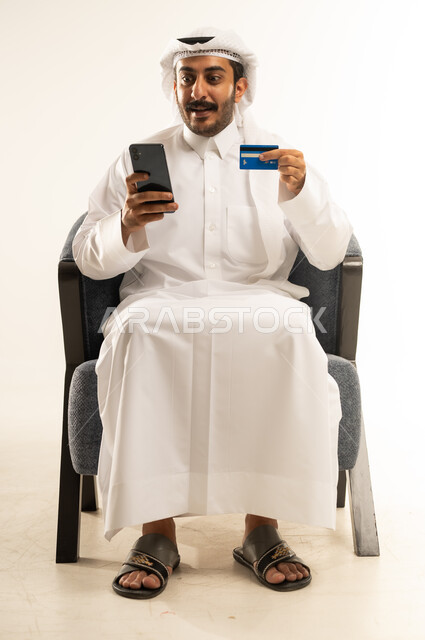 الدفع الالكتروني بالاعتماد على البطاقة البنكية، استخدام التقنيات الحديثة والتكنولوجيا, بورتريه لرجل عربي خليجي قطري يرتدي الثوب التقليدي والغترة يعمل على جهاز الهاتف المحمول، مفهوم التسوق أونلاين، عمليات التبضع والشراء عبر تطبيقات الانترنت، خلفية بيضاء