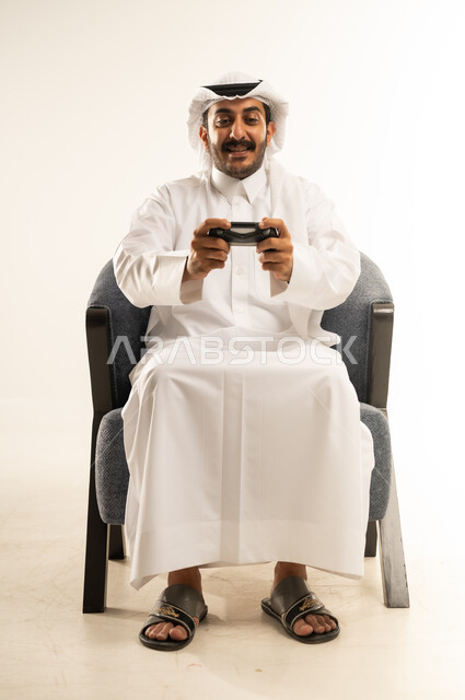 ايماءات السعادة والمتعة باستخدام التقنيات الحديثة، التفاعل والاندماج مع ألعاب الفيديو الترفيهية، بورتريه لرجل عربي خليجي قطري مبتسم يرتدي الثوب والغترة يجلس على كرسي مريح يمسك عصا التحكم بتعابير الاندماج والسرور، خلفية بيضاء
