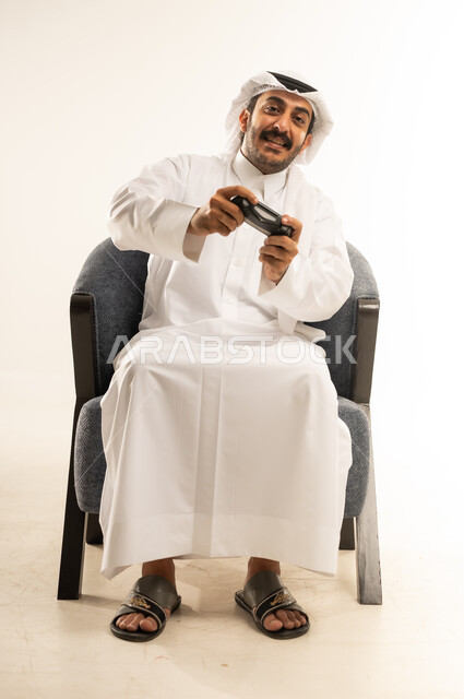 قضاء أوقات ممتعة باستخدام التقنيات الحديثة، التفاعل والاندماج مع ألعاب الفيديو الترفيهية، بورتريه لرجل عربي خليجي قطري مبتسم يرتدي الثوب والغترة يجلس على كرسي مريح يمسك عصا التحكم بتعابير الاندماج والسرور، خلفية بيضاء