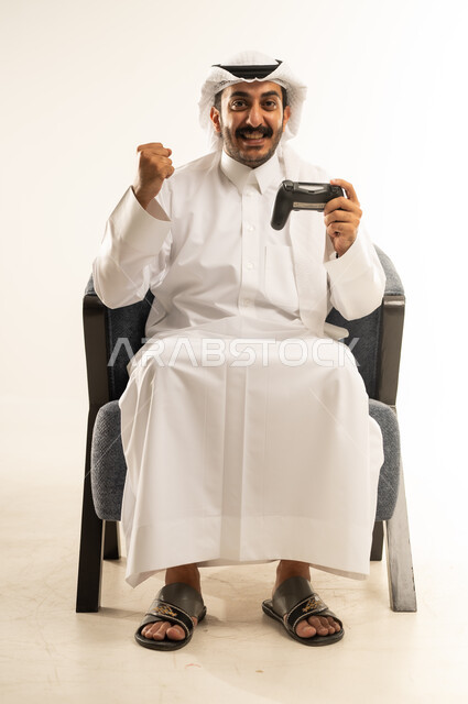 مفهوم الانجاز وتحقيق الاهداف، قضاء أوقات ممتعة باستخدام التقنيات الحديثة، الاندماج والتفاعل في ألعاب الفيديو الترفيهية، بورتريه لرجل عربي خليجي قطري مبتسم يرتدي الثوب والغترة يجلس على كرسي مريح يمسك بيده عصا التحكم يقوم برفع قبضة يده بتعابير الفوز والنجاح، خلفية بيضاء