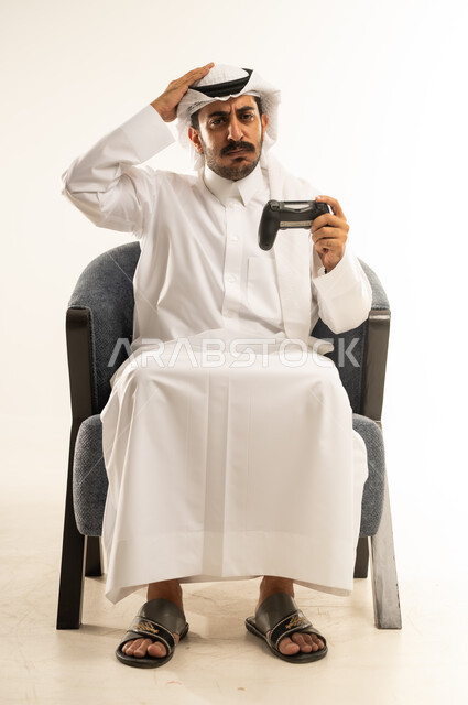 التفاعل والاندماج مع ألعاب الفيديو الترفيهية، بورتريه لرجل عربي خليجي قطري مبتسم يرتدي الثوب والغترة يجلس على كرسي مريح يمسك عصا التحكم يقوم بوضع يده على رأسه بتعابير الخسارة والهزيمة والانزعاج، قضاء أوقات ممتعة باستخدام التقنيات الحديثة، خلفية بيضاء
