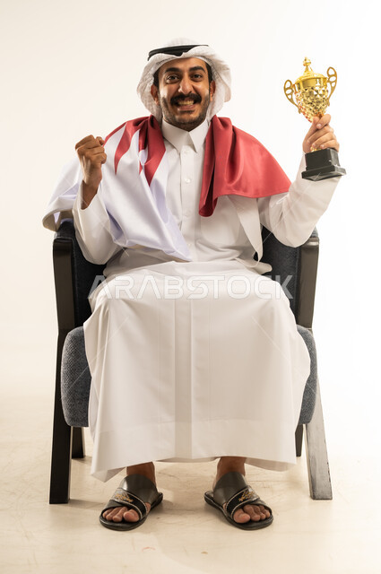 ايماءات النجاح وتحقيق الانجازات، رموز وثقافات وطنية، تشجيع الفرق والمنتخبات, حب الوطن والانتماء اليه، بورتريه لرجل عربي خليجي قطري مبتسم يرتدي الثوب التقليدي والغترة وعلم دولة القطر يجلس على كرسي يحمل بيده كأس البطولة ويرفع قبضة يده بتعابير الفوز, خلفية بيضاء