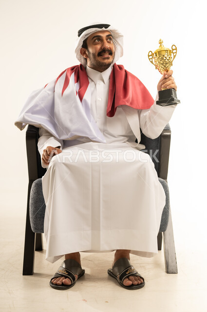 تشجيع الفرق والمنتخبات, حب الوطن والانتماء اليه، بورتريه لرجل عربي خليجي قطري مبتسم يرتدي الثوب التقليدي والغترة وعلم دولة القطر يجلس على كرسي يحمل بيده كأس البطولة بتعابير الفوز, ايماءات النجاح وتحقيق الانجازات، رموز وثقافات وطنية، خلفية بيضاء
