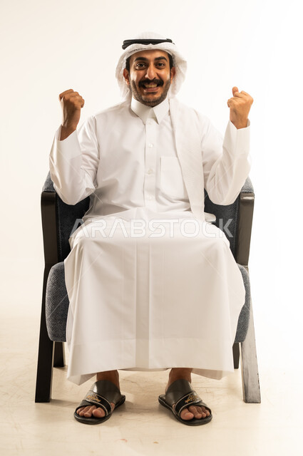 مفهوم الانجاز وتحقيق الاهداف، بورتريه لرجل عربي خليجي قطري مبتسم يرتدي الثوب التقليدي والغترة يجلس على كرسي مريح يرفع قبضة يديه للاعلى بإيماءات الفوز والانتصار والنجاح, النظر الى الكاميرا بتعابير السعادة والقوة والسرور، خلفية بيضاء