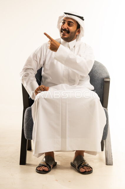 Pointing and explaining something, specifying a specific location, indicating the location of an item, portrait of a Qatari Gulf Arab man wearing a traditional thobe and ghutra sitting on a comfortable chair and pointing with his index finger to a specific target, gestures of happiness and pleasure, white background