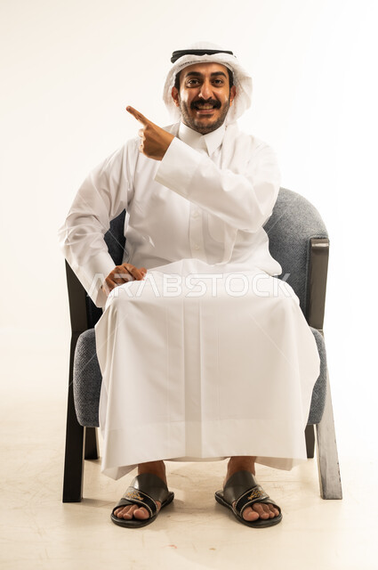 تحديد موقع معين, الدلالة على مكان عنصر، بورتريه لرجل عربي خليجي قطري يرتدي الثوب التقليدي والغترة يجلس ععلى كرسي مريح ويشير بإصبع السبابة إلى هدف محدد، النظر الى الكاميرا بايماءات السعادة والسرور، الإشارة و التوضيح إلى شيء ما, خلفية بيضاء