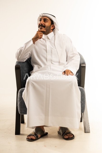 إيماءات التأمل والتمعن والتركيز, بورتريه لرجل عربي خليجي قطري مبتسم يرتدي الثوب التقليدي والغترة يجلس على كرسي يضع يده تحت ذقنه، النظر الى مكان ما بتعابير السعادة، التخطيط لحل مشكلة ما، الشعور بالحيرة وعدم التأكد, خلفية بيضاء