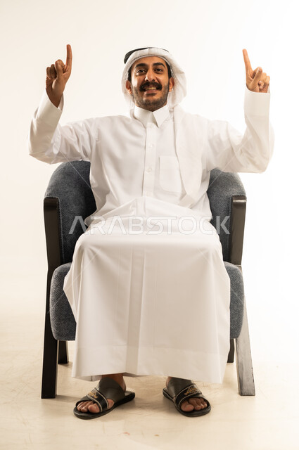 اشارة رقم واحد، النظر إلى الكاميرا بإيماءات الفرح والسرور، بورتريه لشاب عربي خليجي قطري مبتسم يرتدي الثوب التقليدي والغترة يجلس على كرسي مريح يشير بإصبعه السبابة للاعلى إلى شيء محدد بتعابير هذا المكان هنا، خلفية بيضاء .