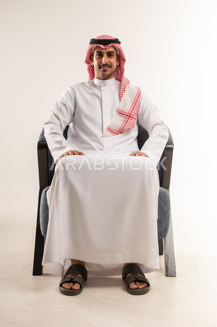 الاهتمام بالمظهر الخارجي والهندام، رجل عربي خليجي بحريني يرتدي الثوب التقليدي والشماغ يجلس على كرسي مريح بتعابير الثقة بالنفس، النظر إلى الكاميرا بإيماءات البشاشة، التقاط الصور الشخصية للوثائق الرسمية، مفهوم الأناقة والشياكة، الجسم بالطول الكامل، خلفية بيضاء